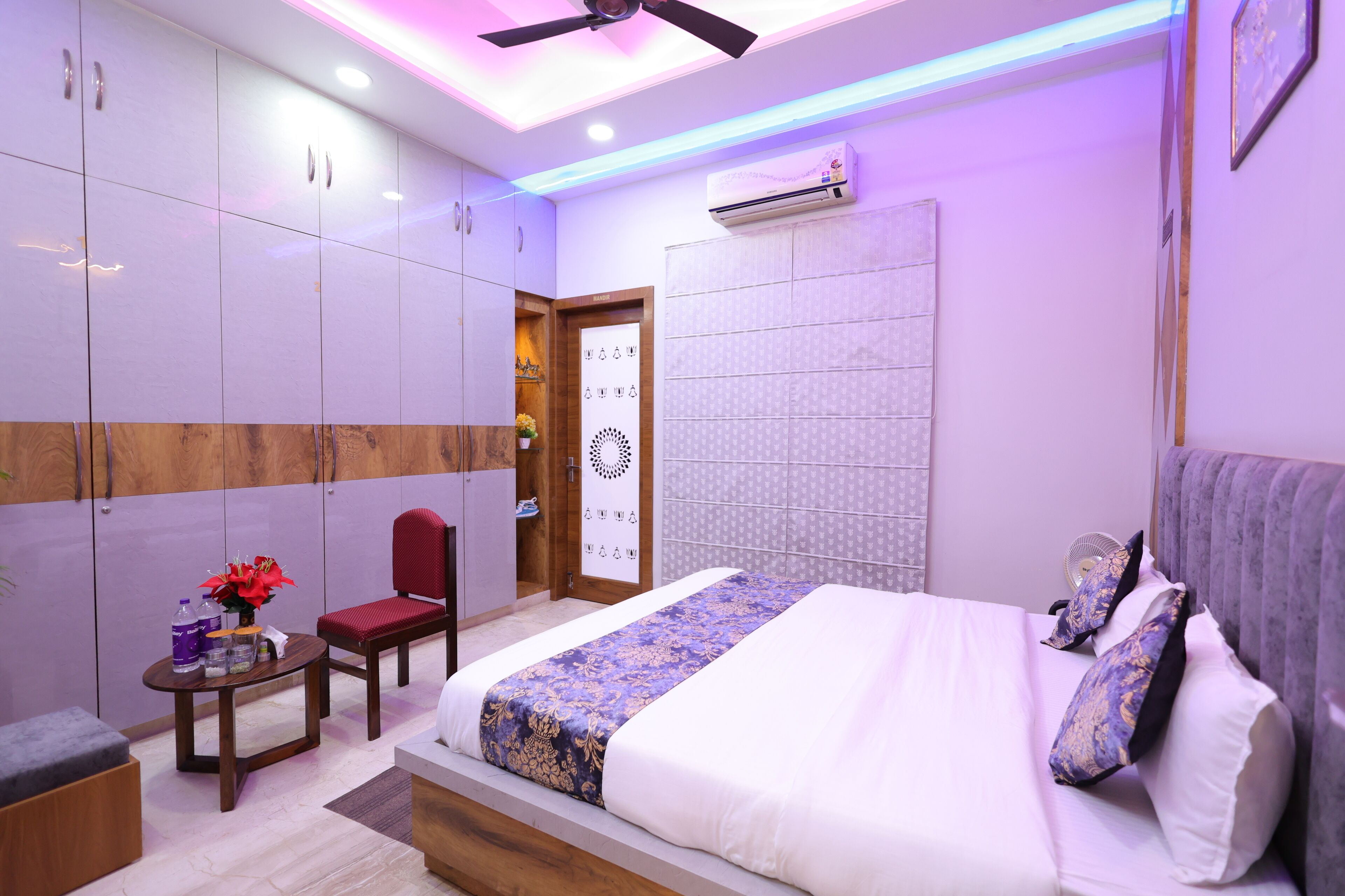 A modern bedroom with a large bed, a desk, and a chair. There is a wardrobe, a door, and a wall-mounted air conditioner.