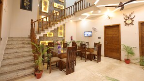 A spacious dining area with a wooden staircase, a ceiling fan, and a television on the wall. - Amritam Palace A Luxury AC Home Stay (Shri Madhopur)