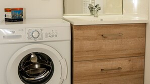 A compact laundry area with a washing machine, a wooden cabinet with drawers, a mirror, and a small shelf with a box. - Confortable T2 avec terrasse, parking & vue étang (Sète)