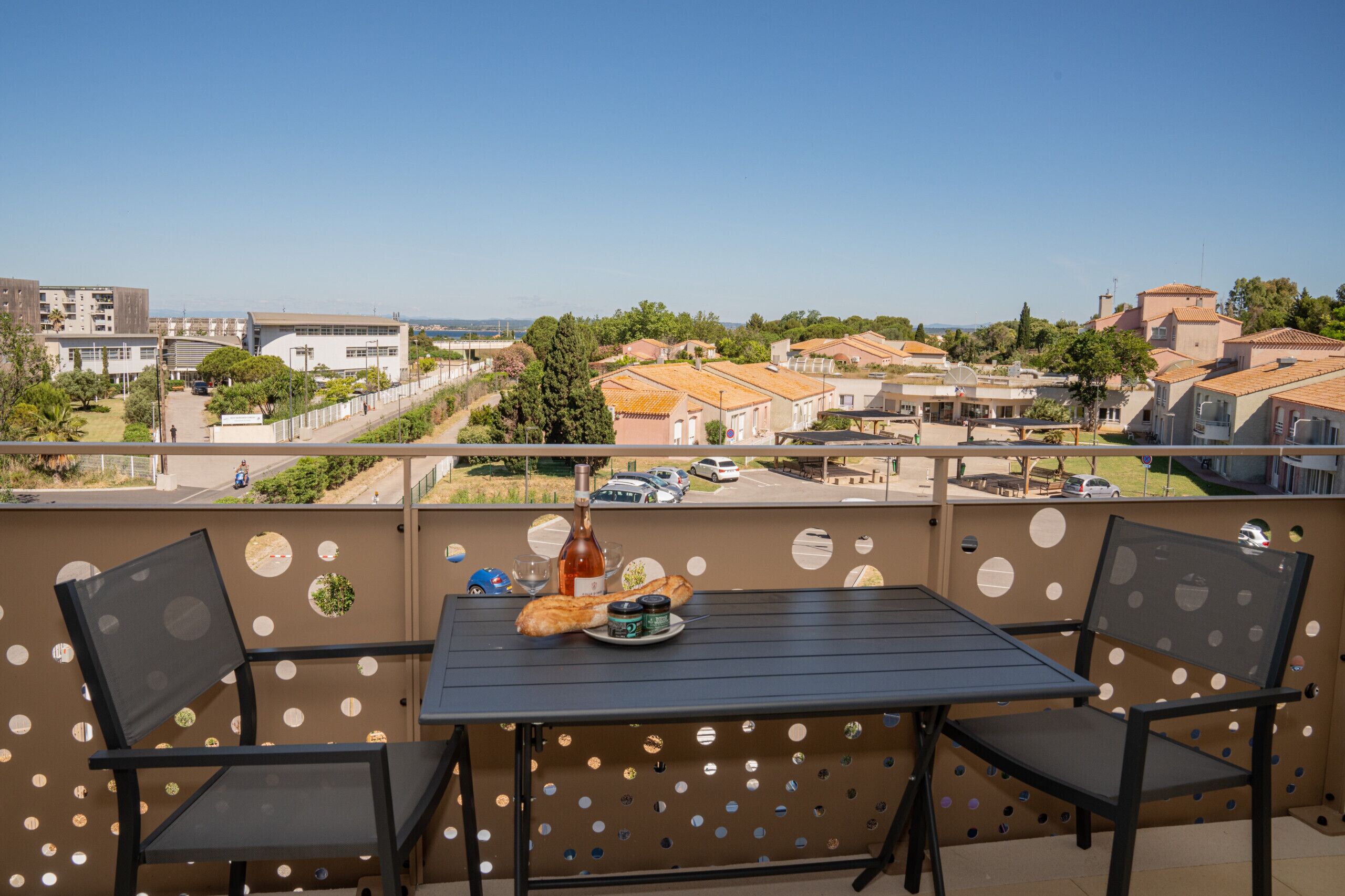 A balcony with a table and chairs, a bottle of wine, and a plate with food. The balcony overlooks a residential area with houses and a road.