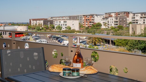 A balcony with a table set for a meal, a bottle, and two glasses. - Confortable T2 avec terrasse, parking & vue étang (Sète)
