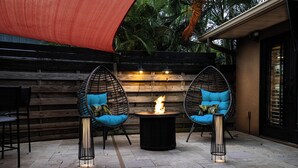 An outdoor patio with two wicker chairs, a round table with a fire, and a wooden wall in the background. - Heated Pool | Game Room | Arcade | Sleeps14 | BBQ (Bradenton)