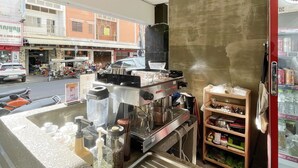 Ett kafé med en kaffemaskin, ett kylskåp fyllt med Coca-Cola och utsikt över gatan med parkerade bilar och byggnader.