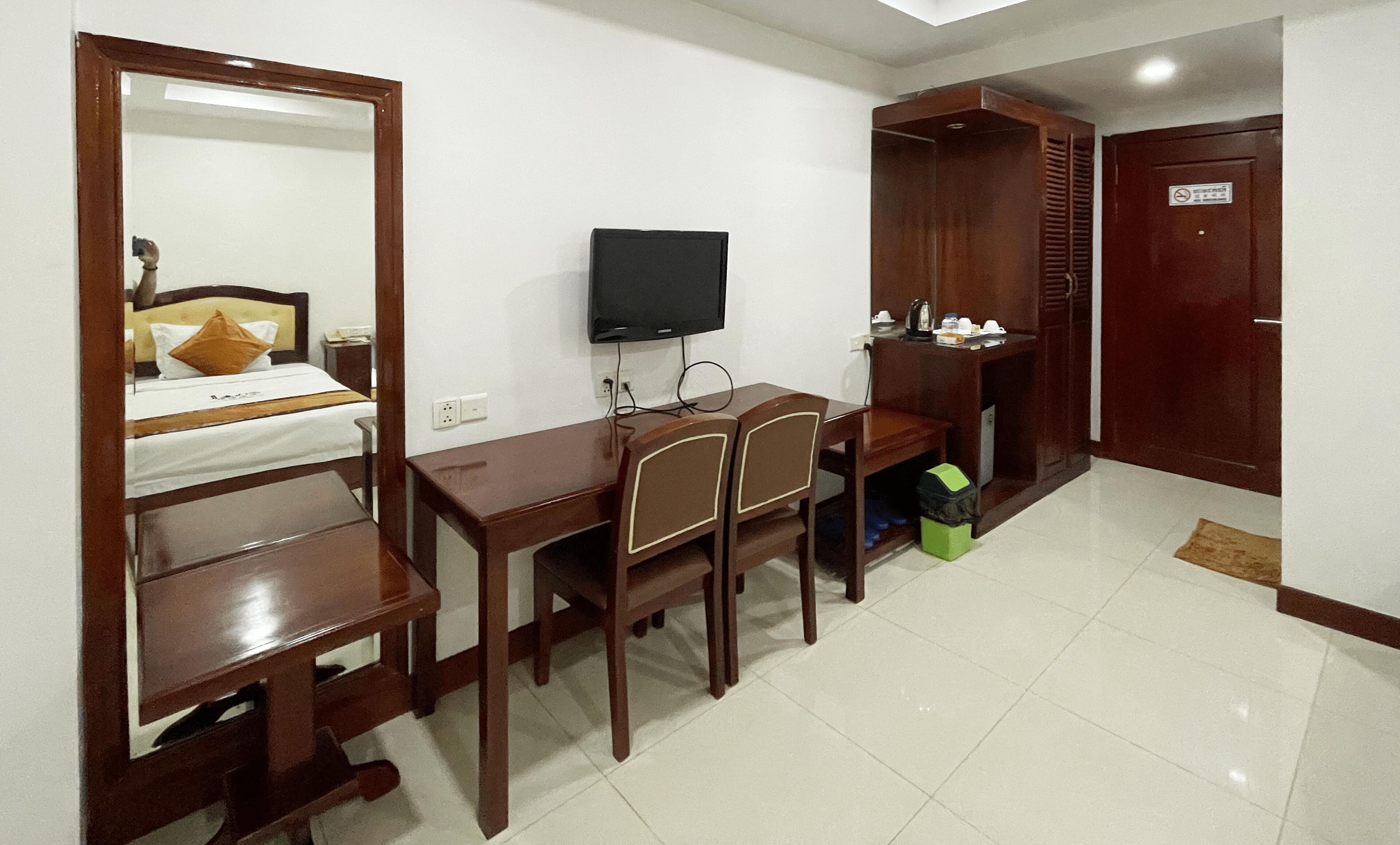 A hotel room with a bed, a desk with chairs, a television, a wardrobe, and a door with a sign.