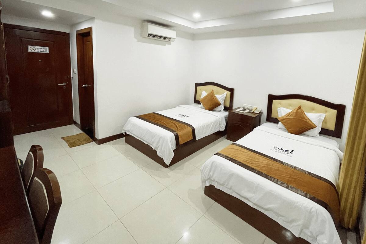 A hotel room with two beds, a wooden door, an air conditioning unit, and a bedside table with a phone.