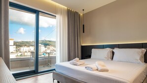 A modern hotel room with a large bed, a balcony with a city view, and a large window. - Sasso Hotel (Vlorë)