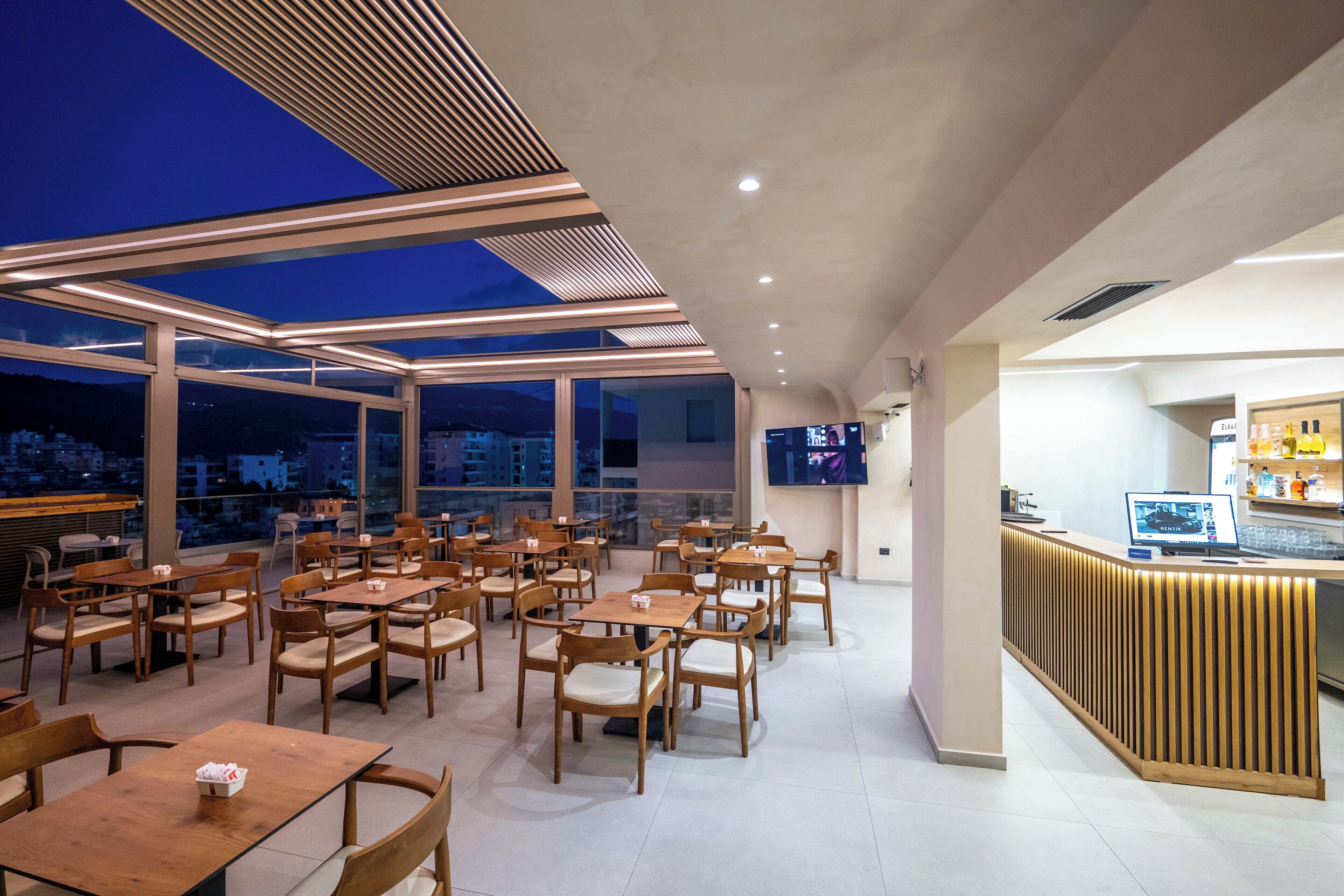 A modern restaurant with wooden tables and chairs, a bar area, and large windows offering a city view.