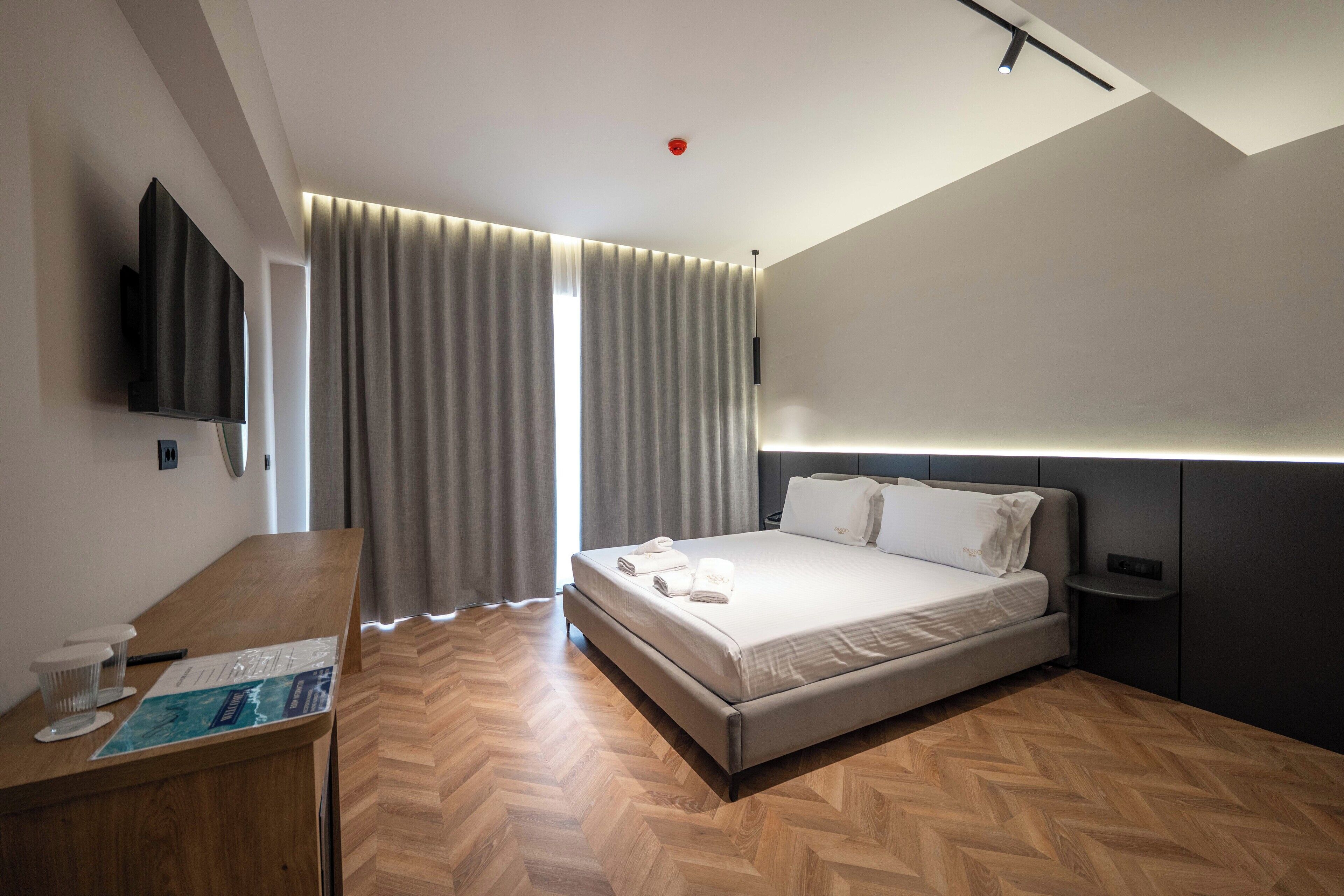 A modern hotel room with a large bed, a desk with a chair, a flat-screen TV mounted on the wall, and a large window with curtains.