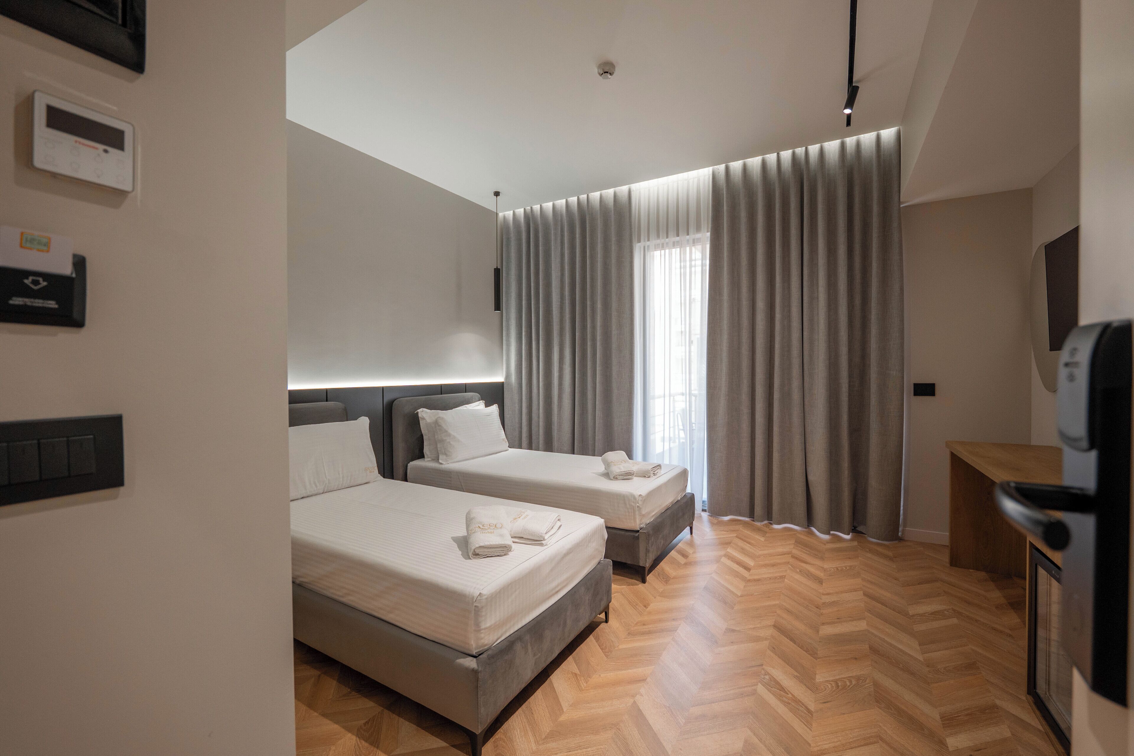 Une chambre d’hôtel avec deux lits, un parquet en bois disposé en point de Hongrie, une grande fenêtre avec des rideaux, un téléviseur intégré et un panneau de commande mural.