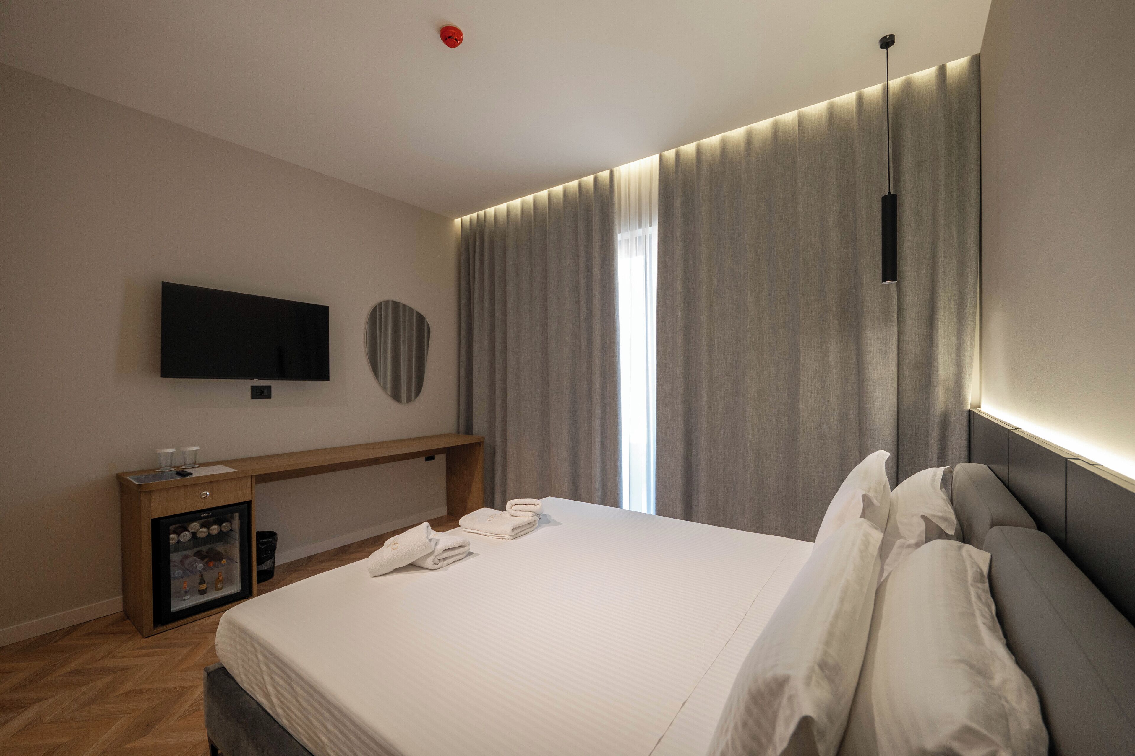 Une chambre d’hôtel moderne dotée d’un grand lit, d’un téléviseur à écran plat fixé au mur, d’un minibar et d’une grande fenêtre avec des rideaux.