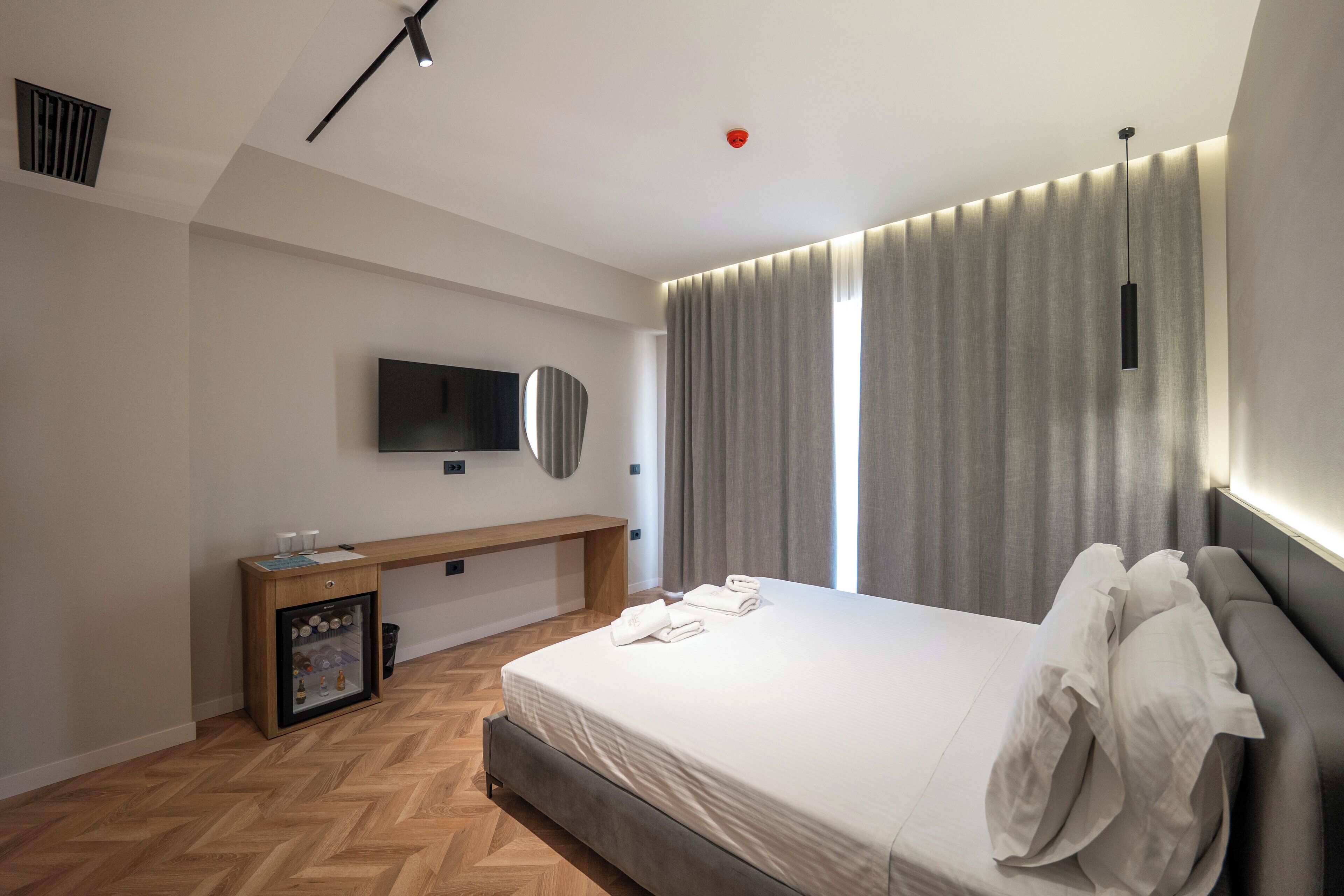 A modern hotel room with a large bed, a bedside table, a TV mounted on the wall, and a window with curtains.