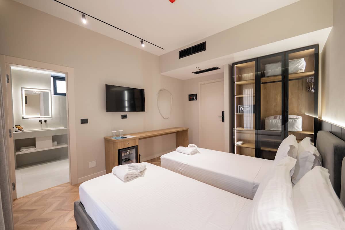 A modern hotel room with two beds, a TV, a minibar, a wooden desk, and a walk-in closet.