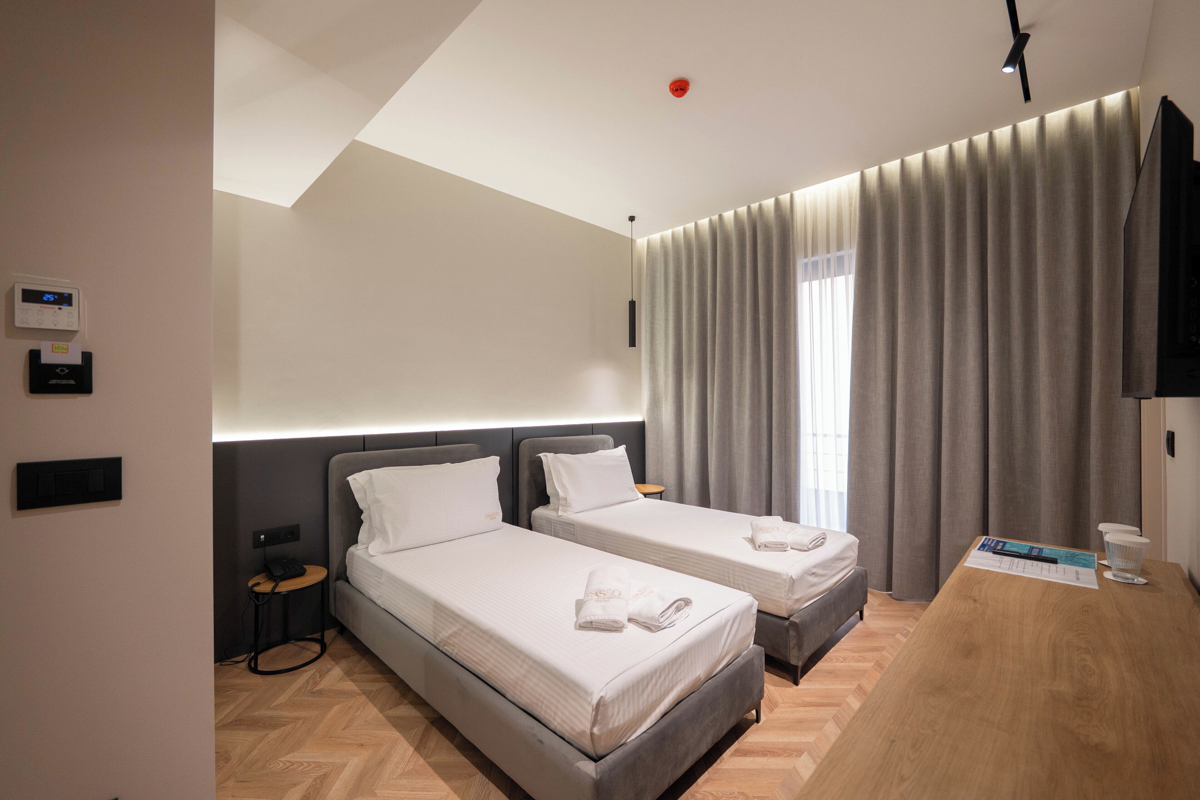 A modern hotel room with two beds, a wooden desk, a flat-screen TV, and a large window with curtains.