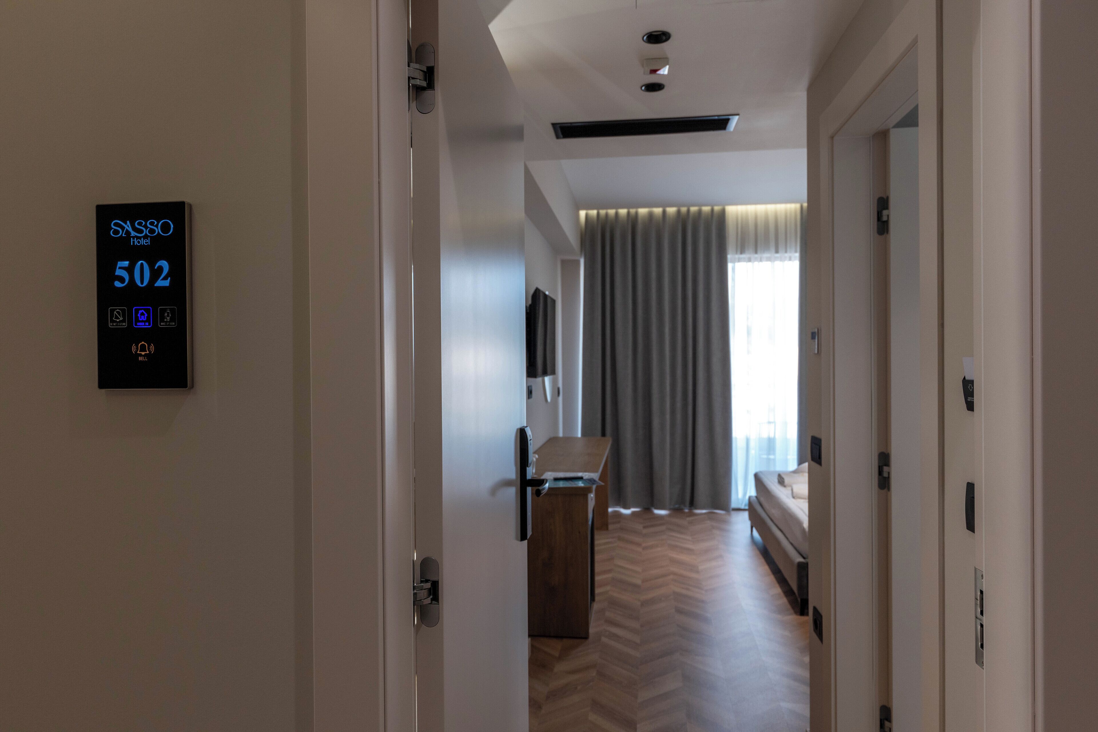 A hotel room with a digital display showing room number 502, a bed, a desk, and a large window with curtains.