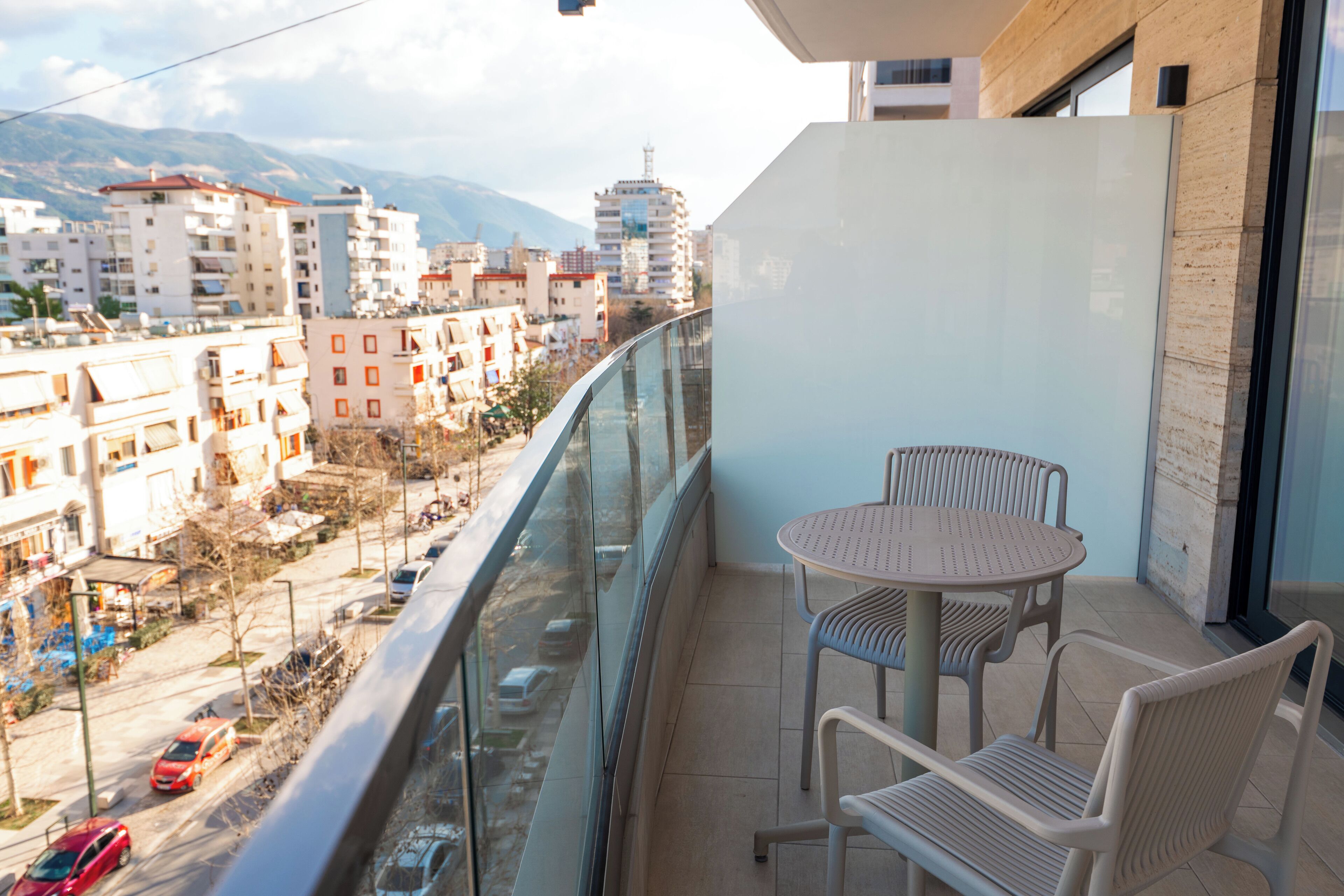 A balcony with a glass railing overlooking a street with parked cars and buildings. There is a round table with two chairs.