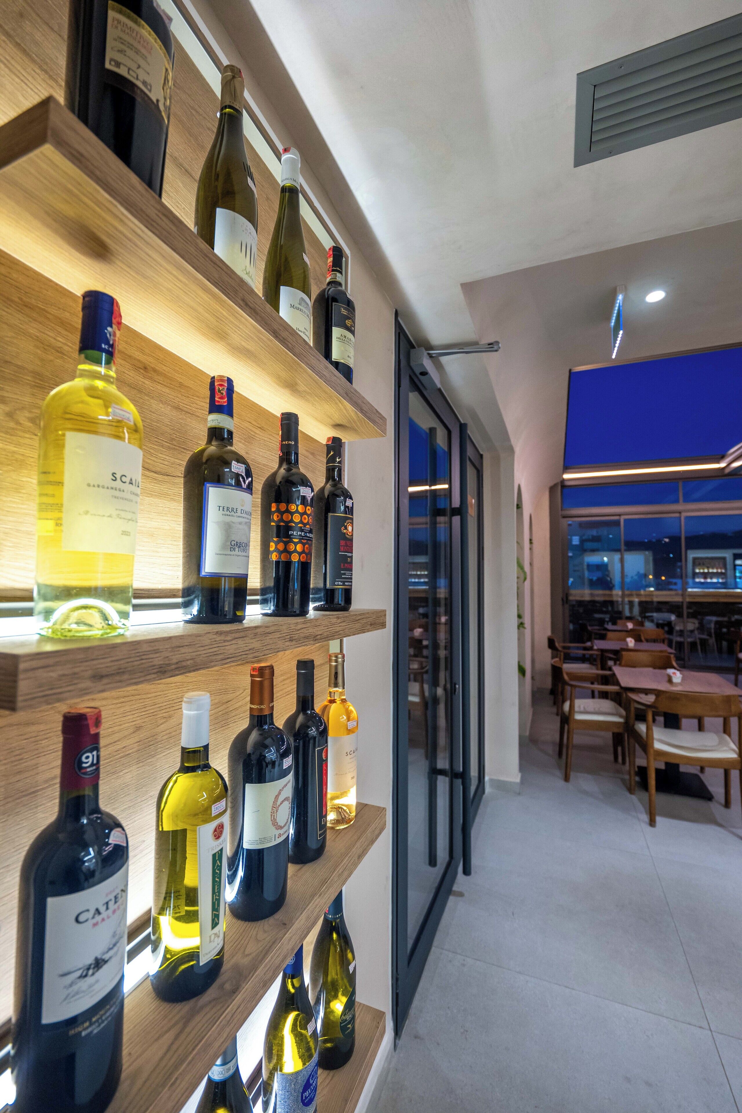 A wine cellar with shelves of various wine bottles, a glass door leading to a dining area, and a view of the city at dusk.