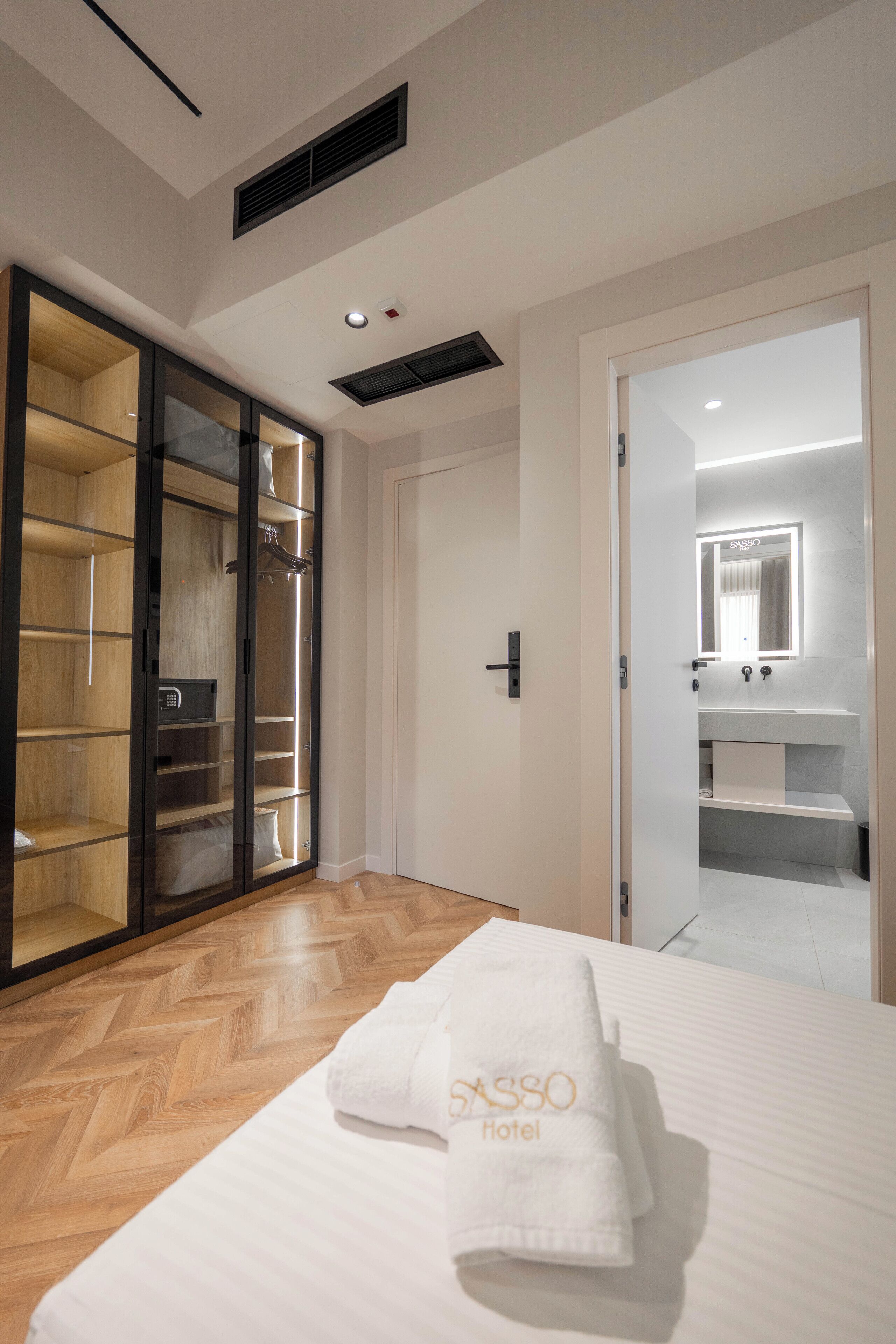 Ein Hotelzimmer mit großem Kleiderschrank, einem Bett mit weißem Handtuch und einem durchgegangenen Türflügel einsehbarer Badezimmer.