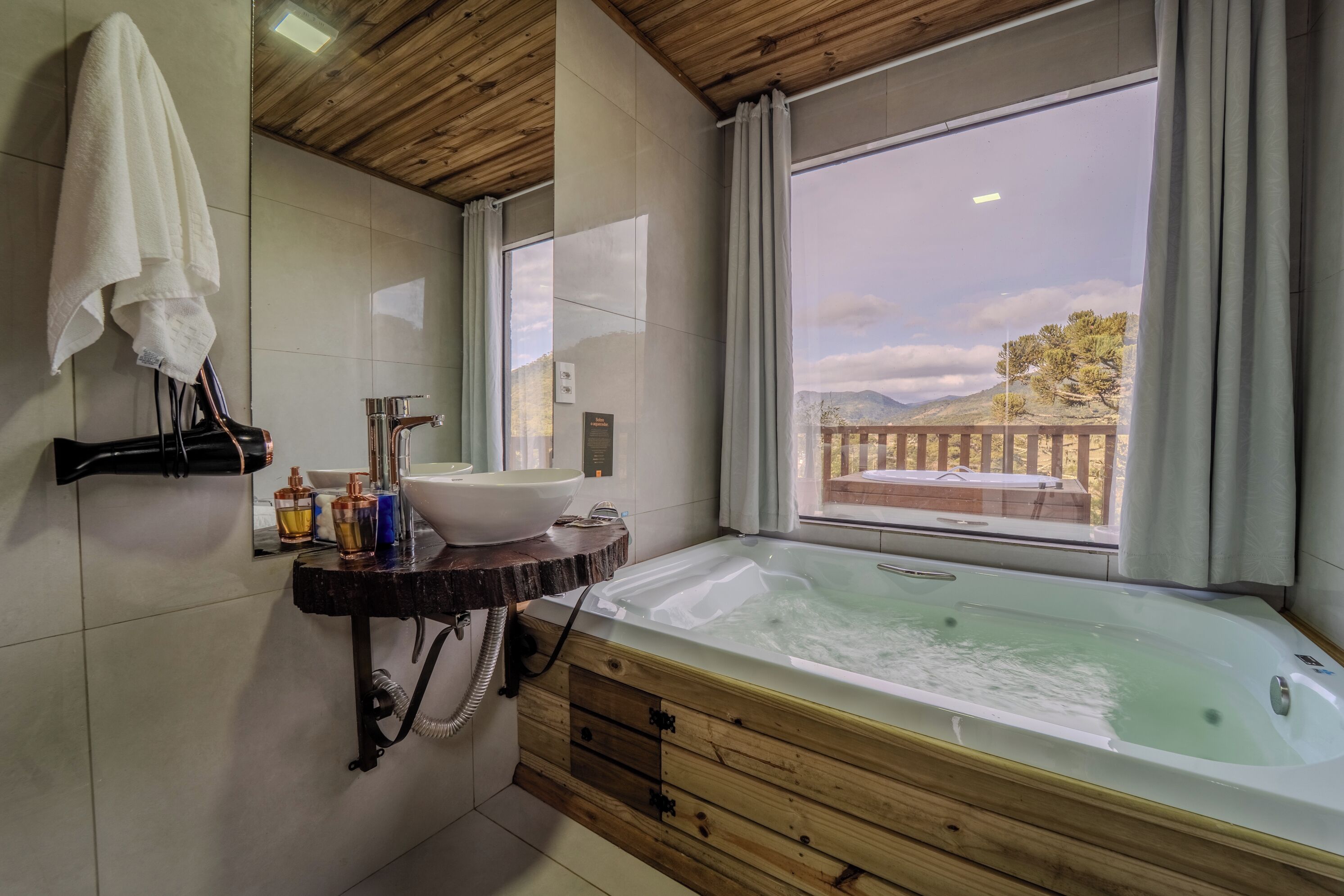 Un baño con tocador de madera, una bañera grande, un lavabo y una ventana con vistas a un balcón y montañas.