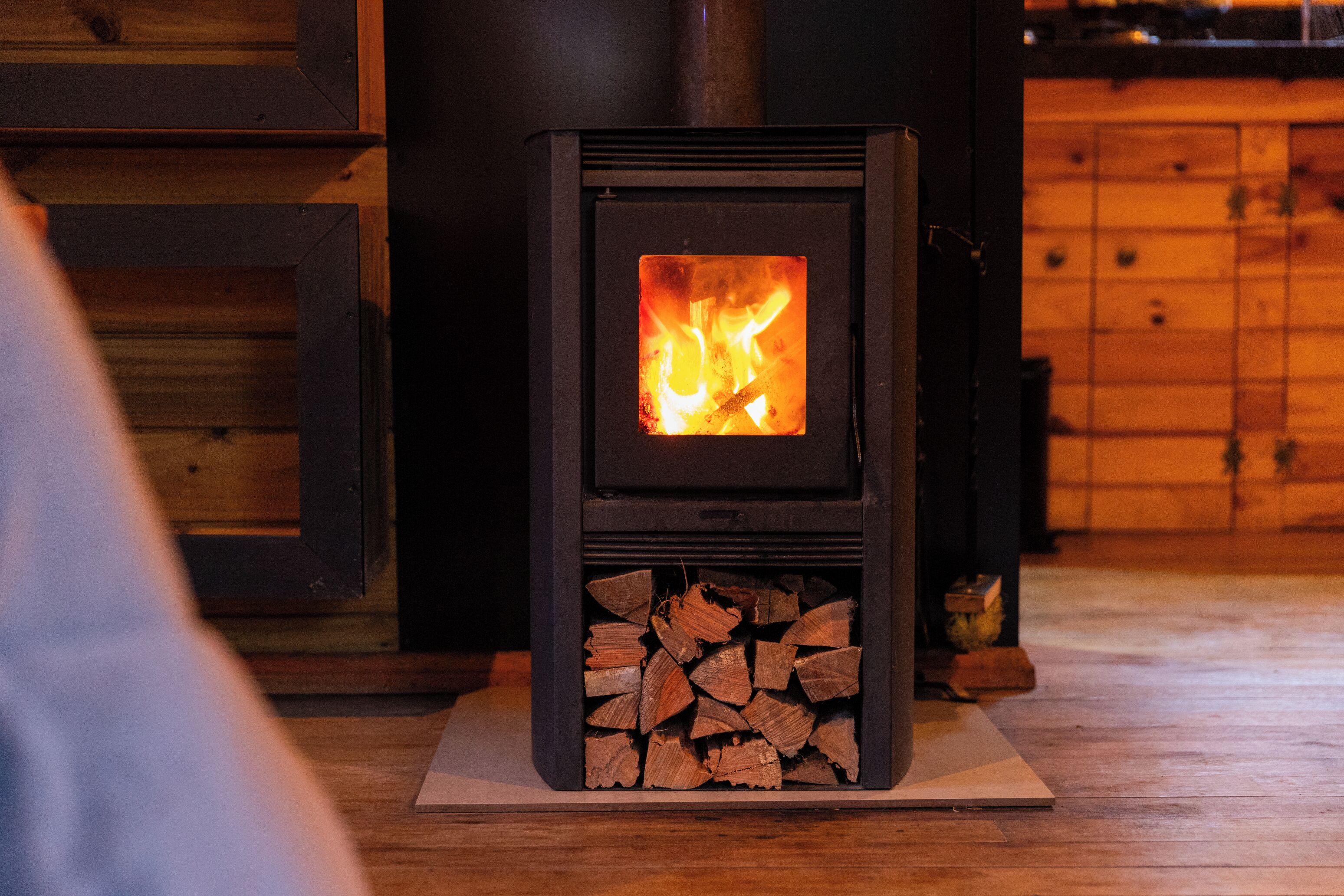 A wood-burning stove with visible flames, a stack of firewood, and a wooden wall in the background.