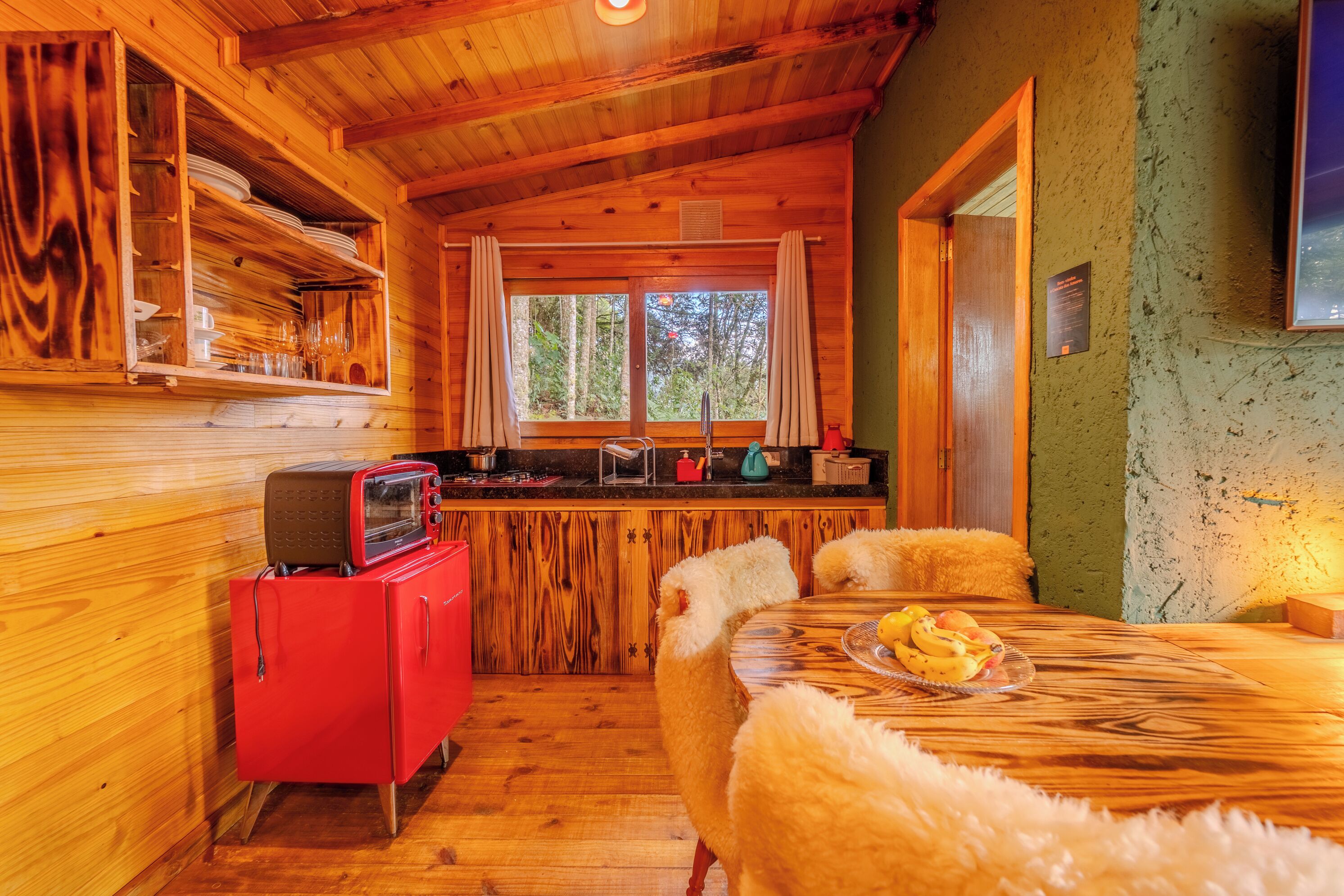 A wooden cabin interior with a dining area, a kitchen with a red appliance, and a window with curtains.