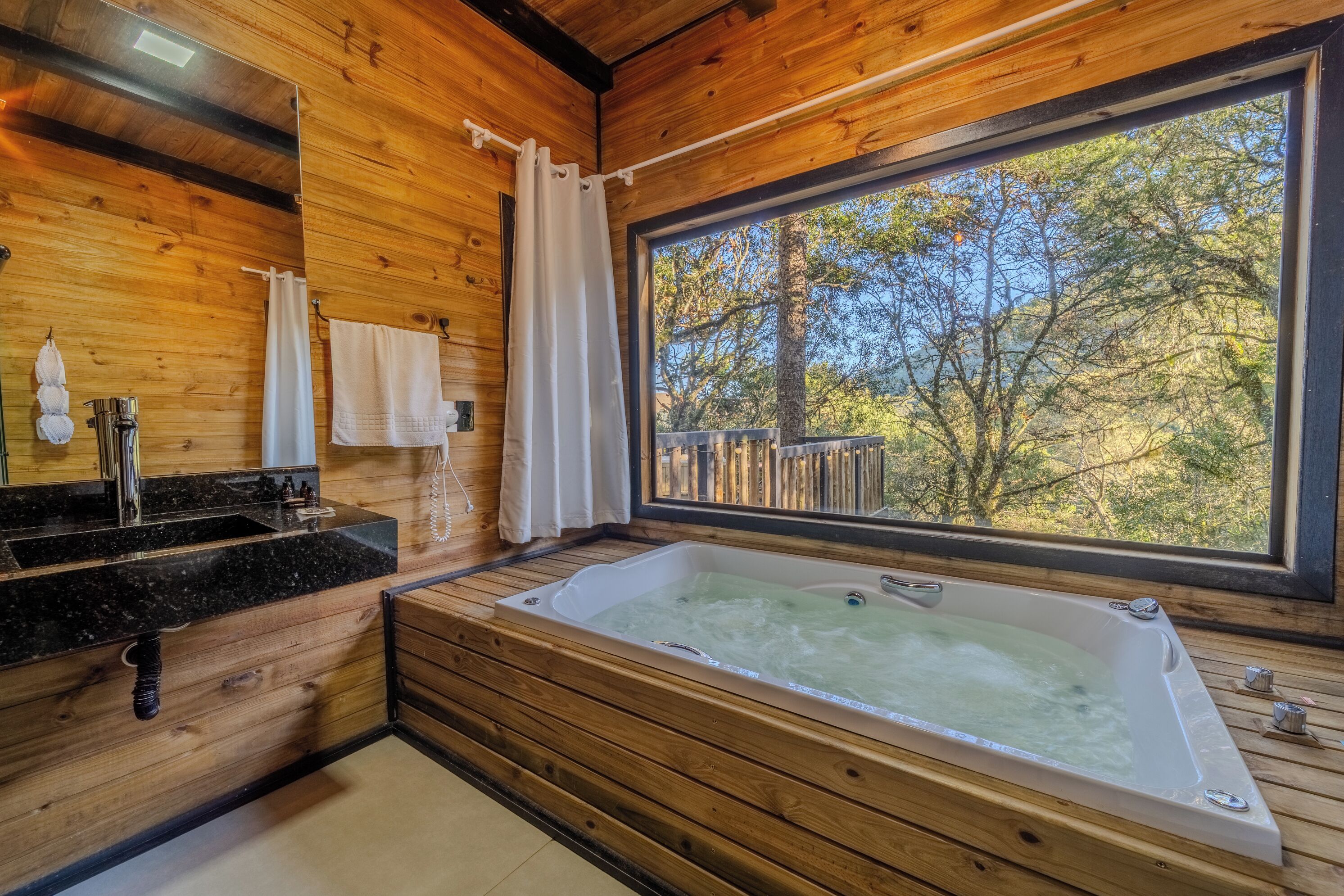A wooden cabin bathroom with a large window, a bathtub, a sink, and a mirror.