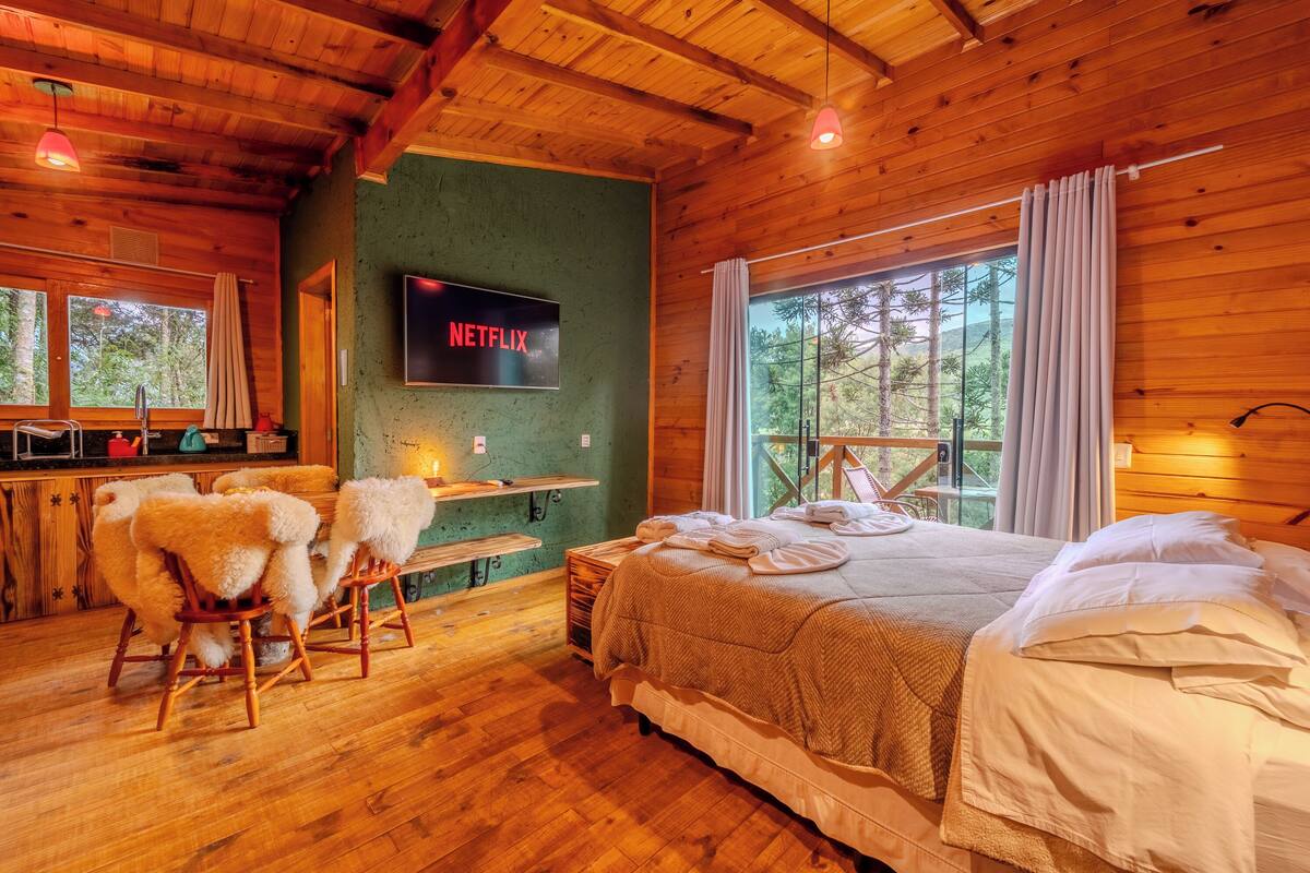 Une pièce de style cabane en bois, avec un lit, un téléviseur fixé au mur, un banc en bois et une fenêtre donnant sur la forêt.