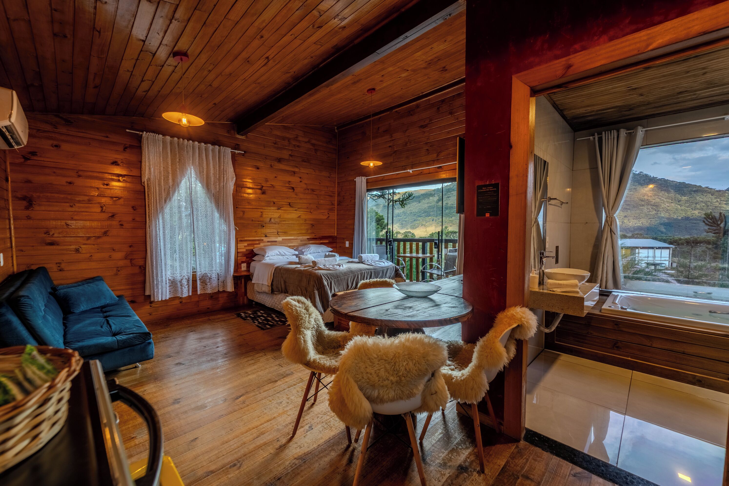 Interior acogedor de una cabaña de madera con cama, una mesa redonda de comedor, sillas mullidas y vistas a un paisaje montañoso a través de un ventanal grande.