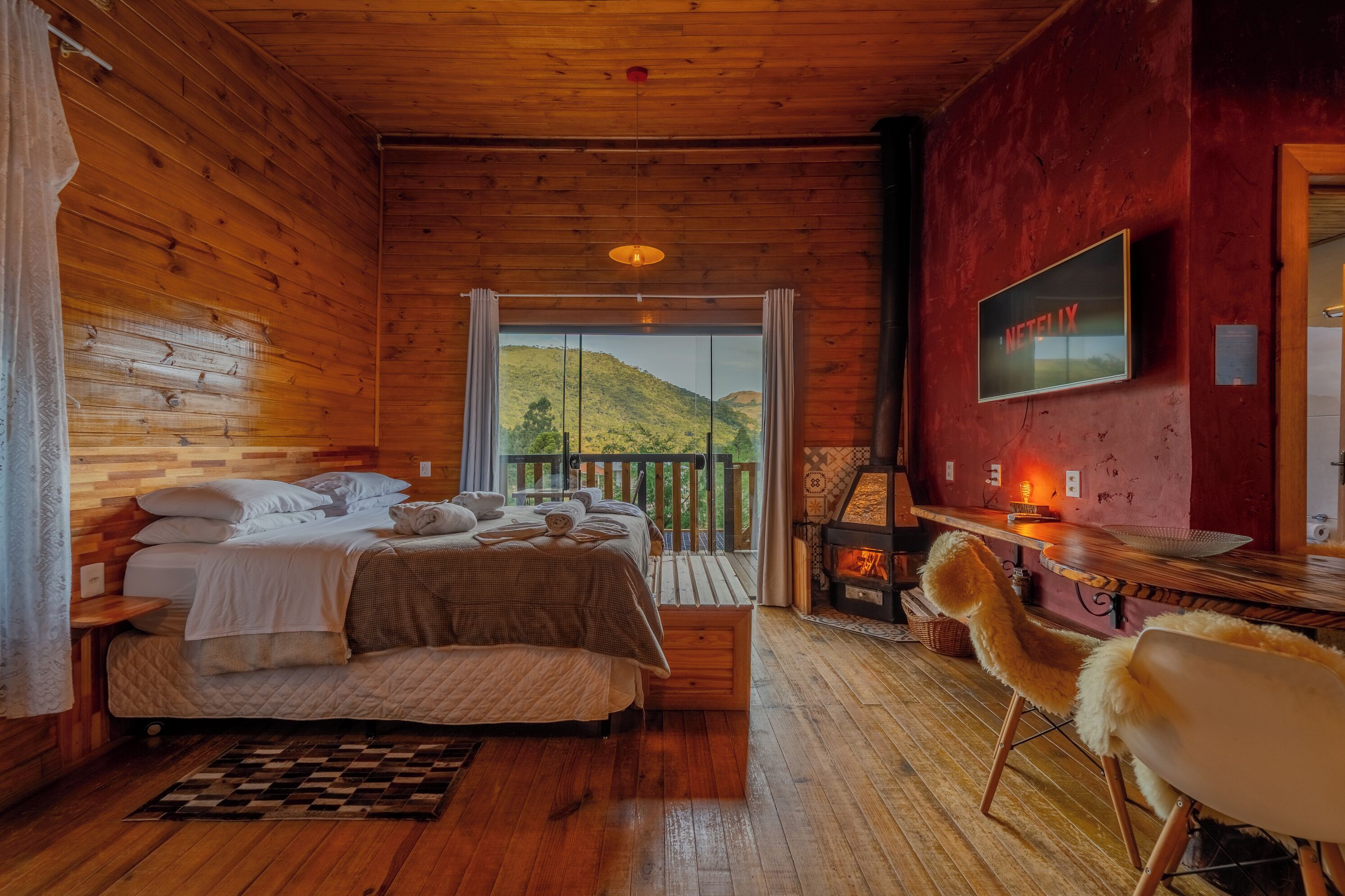 A cozy wooden cabin bedroom with a bed, a TV, a fireplace, and a view of the outdoors.