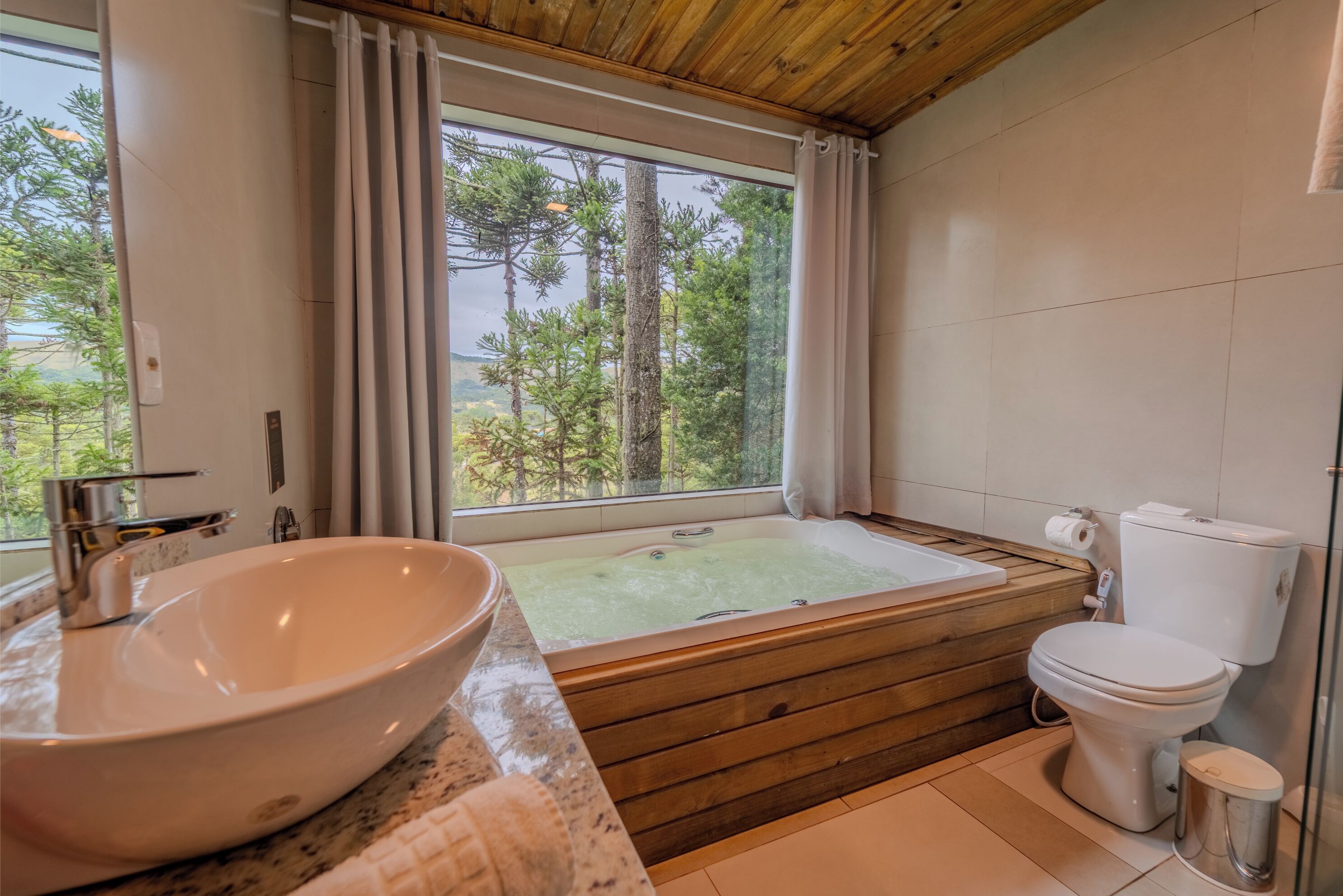 Een moderne badkamer met een groot raam, een houten ligbad, een wit toilet en een groot wastafelbak.