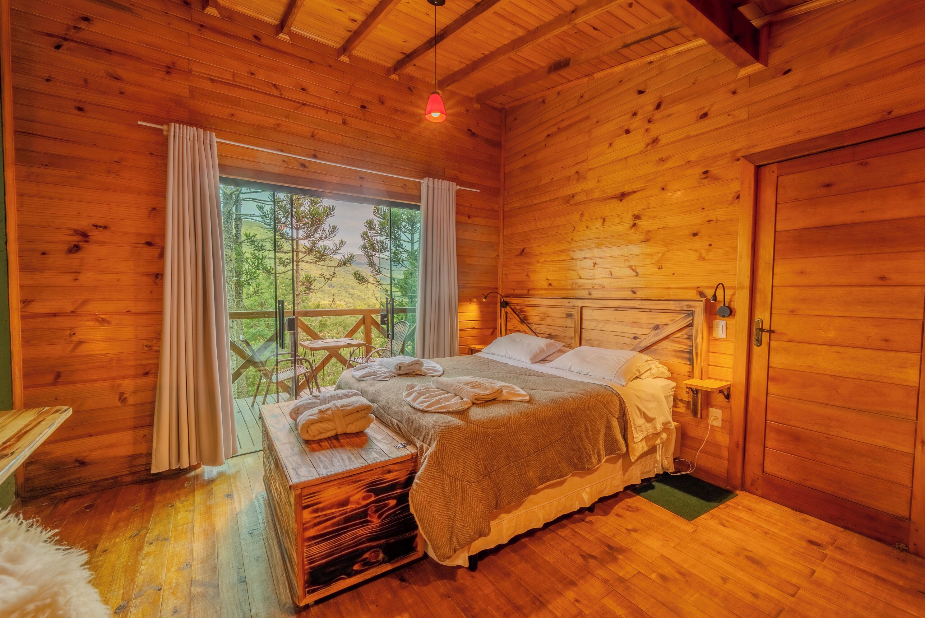 A wooden cabin bedroom with a bed, a small table with towels, a window with curtains, and a view of trees.