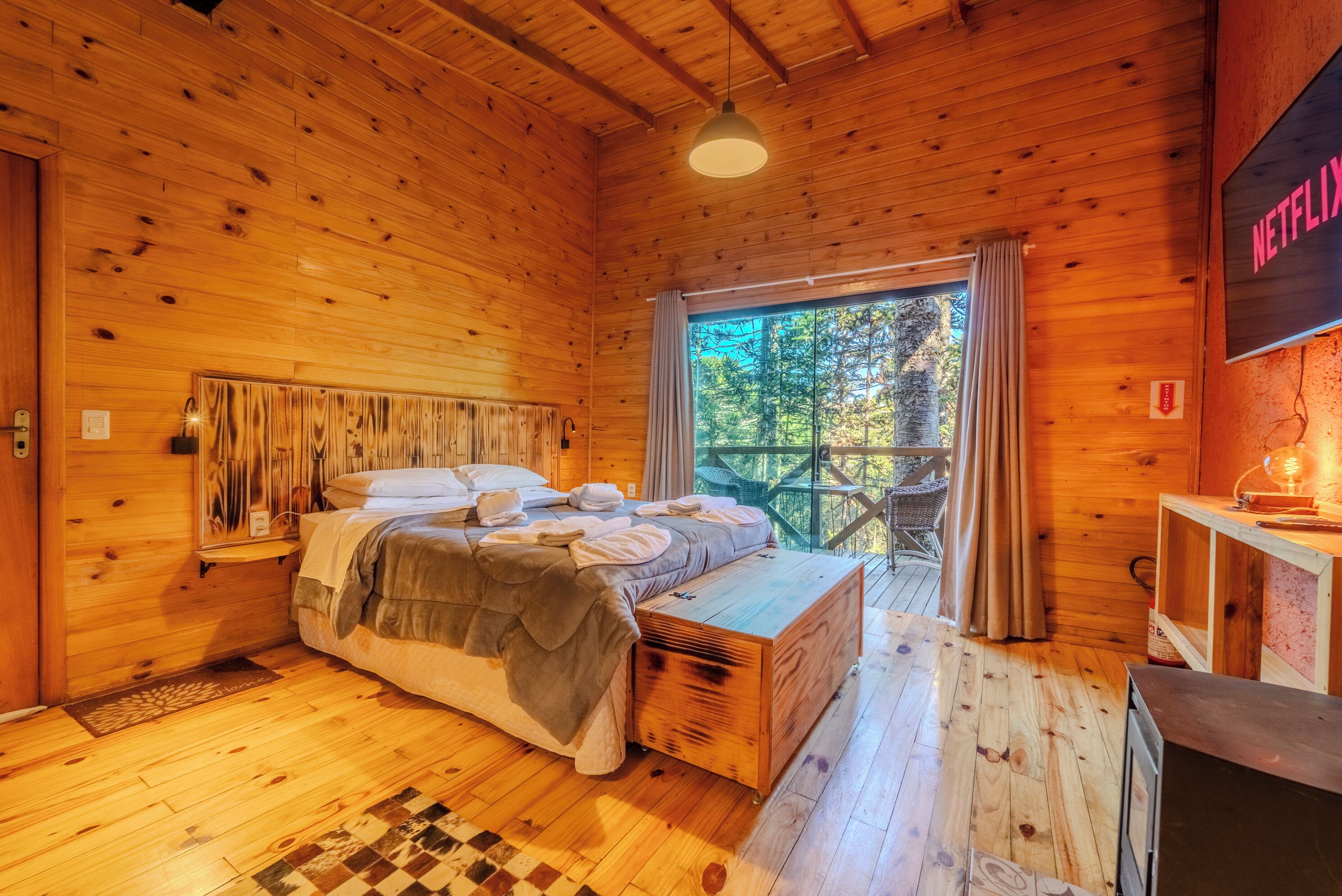 Une chambre aménagée dans une cabane en bois, avec un lit, un banc, une télévision et une vue sur l’extérieur.