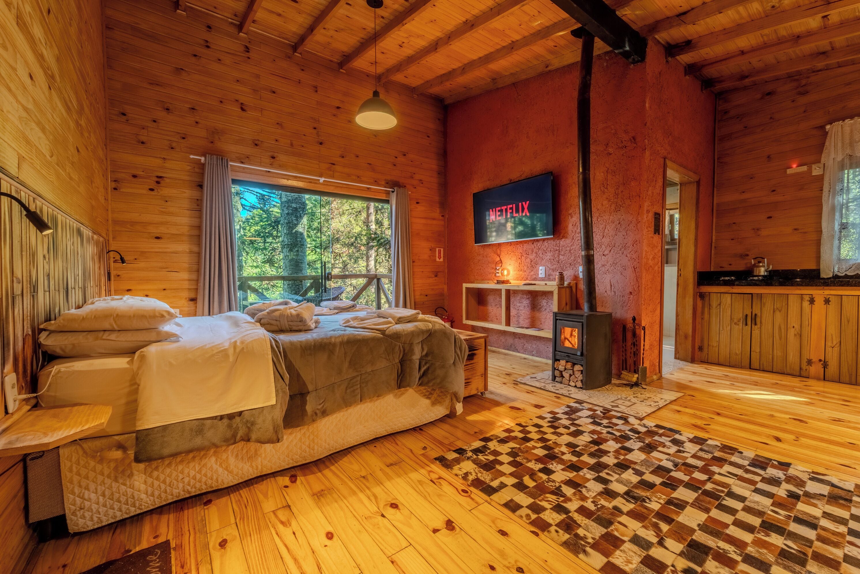 Una habitación acogedora tipo cabaña con una cama, un televisor montado en la pared y una ventana con vista a un bosque.