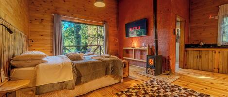 A cozy cabin room with a bed, a television mounted on the wall, and a window overlooking a forest.