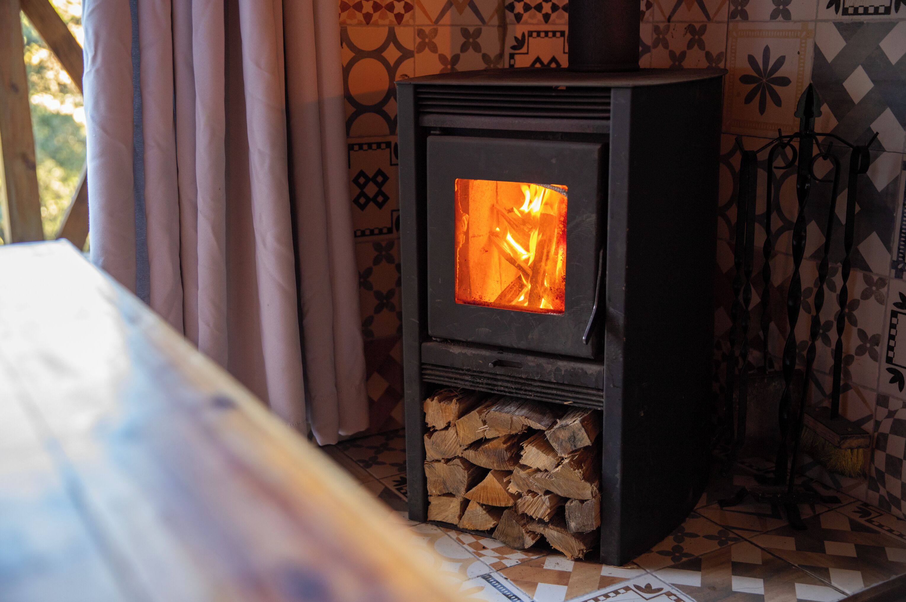A wood-burning stove with visible fire, a bed, and patterned tiles in the background.