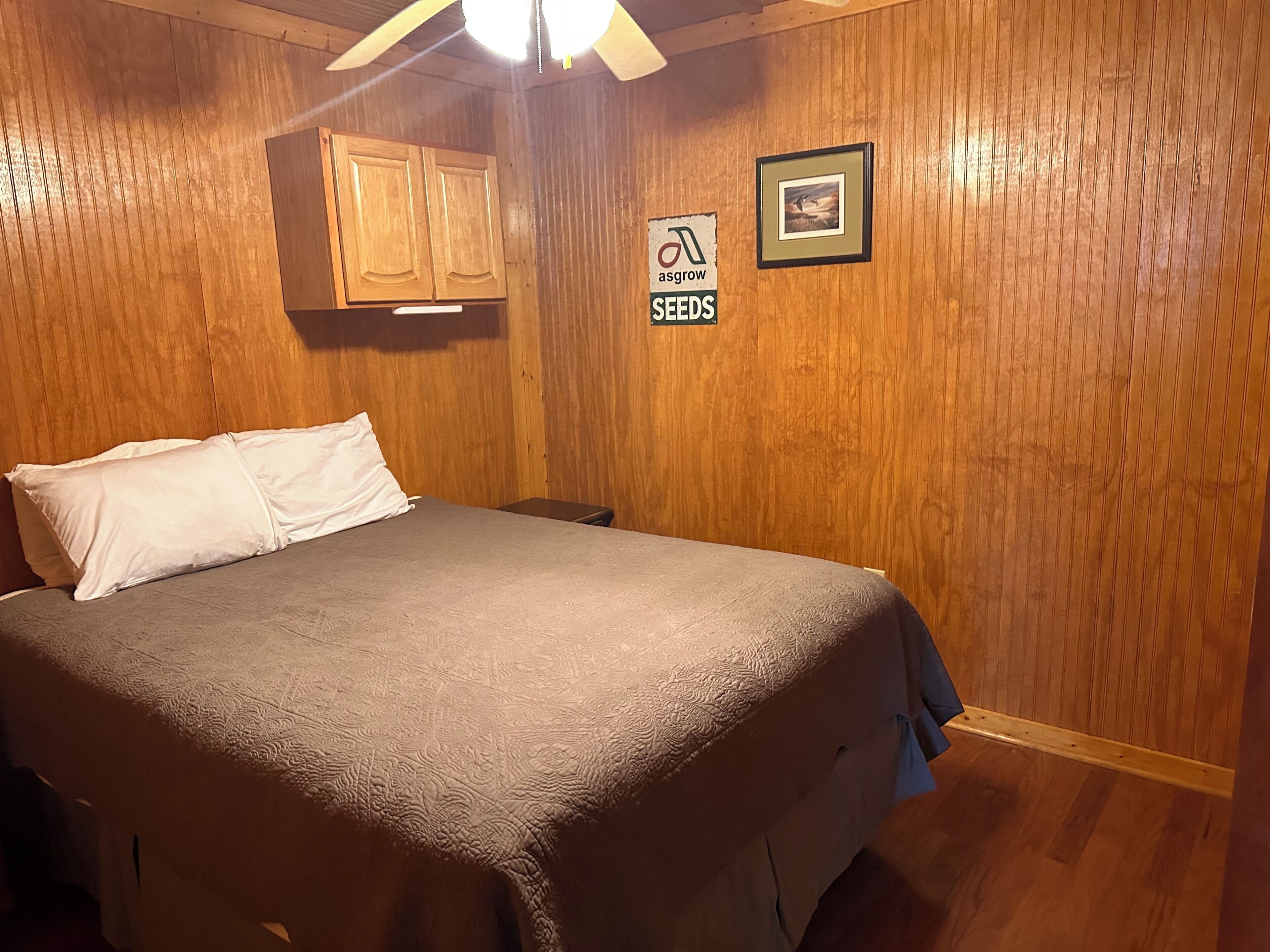 A wooden-paneled room with a bed, a ceiling fan, a framed picture, and a sign on the wall.
