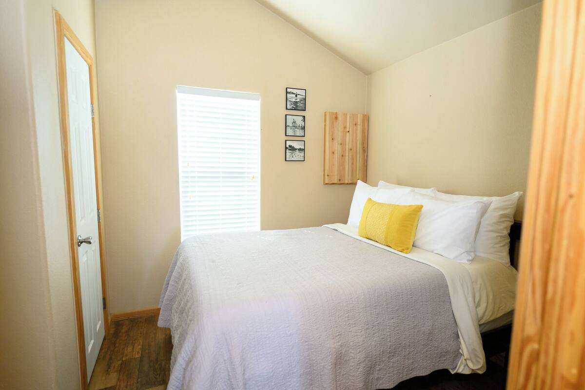 A bedroom with a bed, a window with blinds, and three framed pictures on the wall.