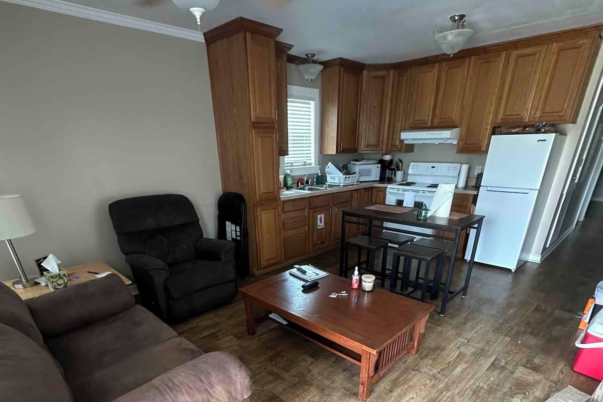A living room with a brown leather sofa, a wooden coffee table, and a black armchair. There is a kitchen area with wooden cabinets, a white refrigerator, and a microwave.