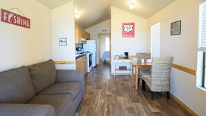A compact living space with a sofa, dining table, wicker chairs, a kitchenette, and a hallway leading to a bedroom. - Red Leaf Cottages (Lake Village)