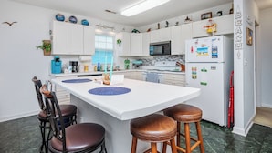 A modern kitchen with white cabinets, a central island with stools, a refrigerator with magnets, and a window with blinds.