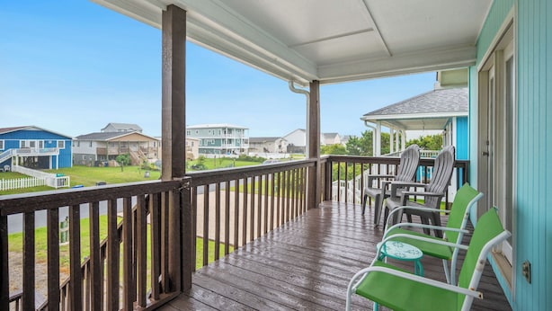A covered porch with wooden railings and green chairs, overlooking a residential area with houses and a grassy lawn.