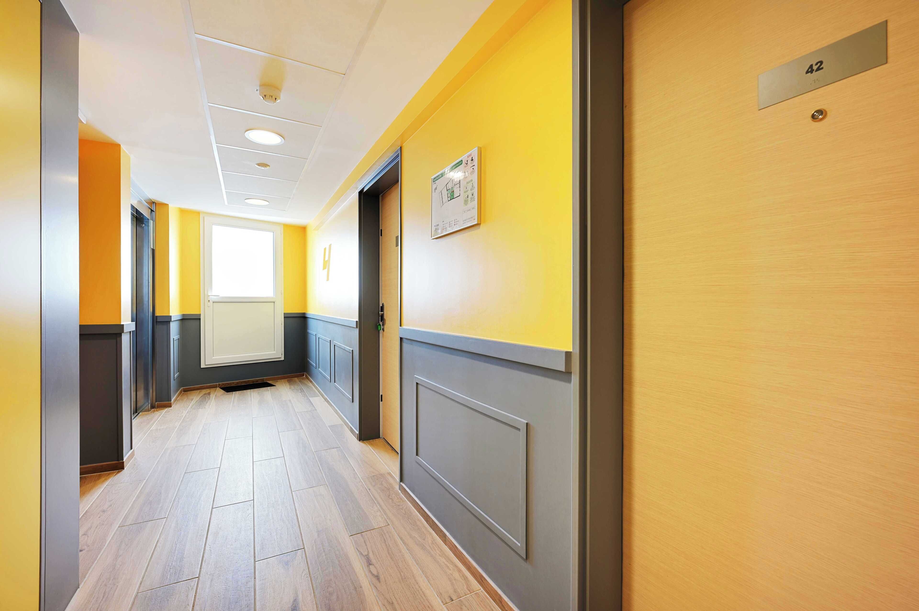 A hallway with a yellow wall, a door with a number plate, and a window at the end.