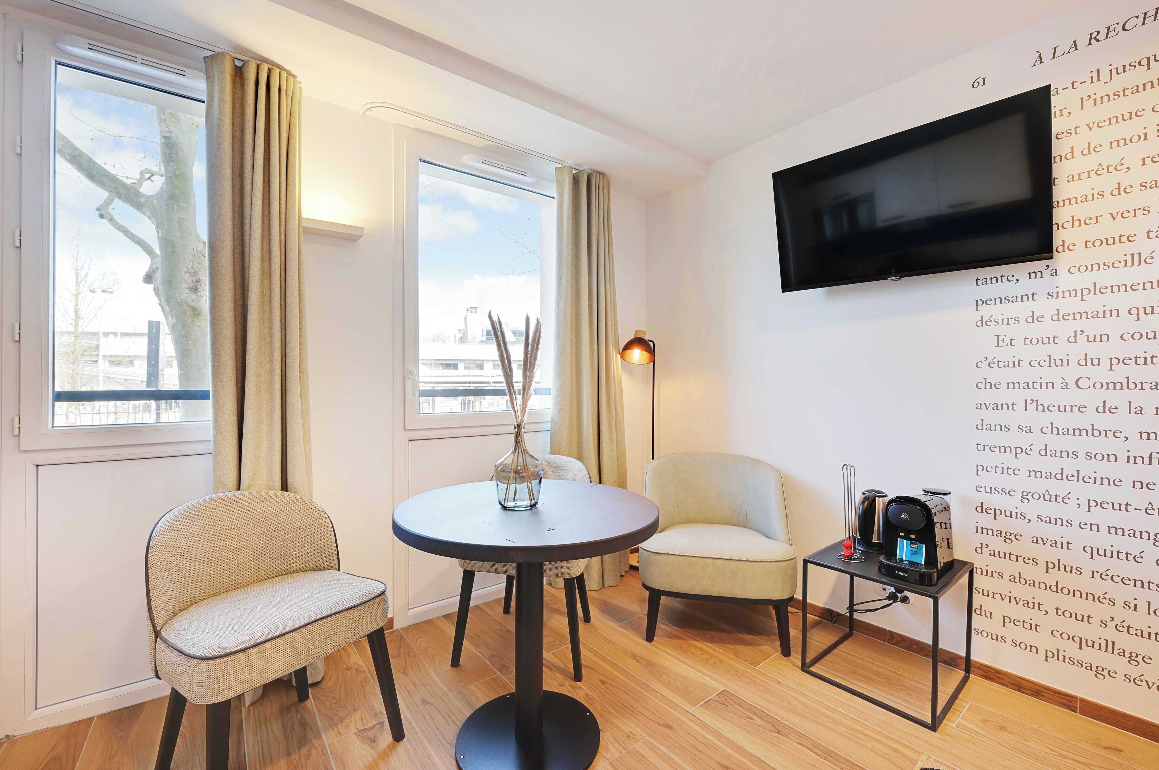 A room with a round table, two chairs, a TV mounted on the wall, and a small table with a lamp and a coffee machine.