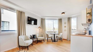 A modern living room with a flat-screen TV mounted on a brick wall, a round dining table with chairs, a sofa, and a coffee table. - Résidence du Parc Vanves (Vanves)
