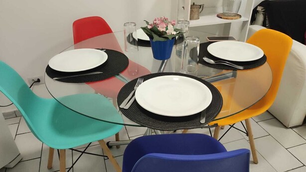 A dining area with a glass table, four chairs in different colors, and a white plate on each chair. - Magnifico Apartamento Central (Curitiba)