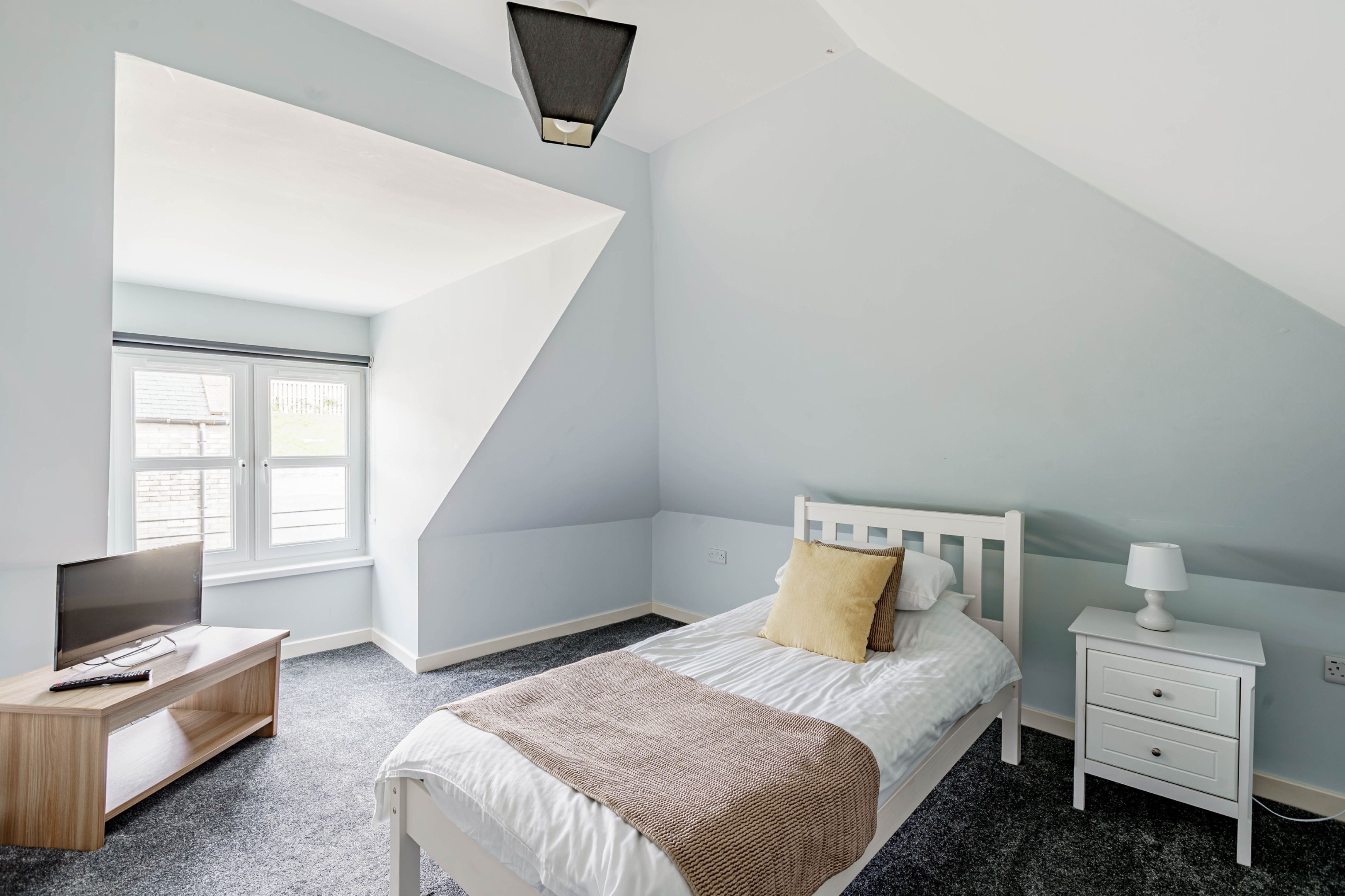 Un dormitorio con cama, mesita de noche, lámpara y televisor sobre un soporte.