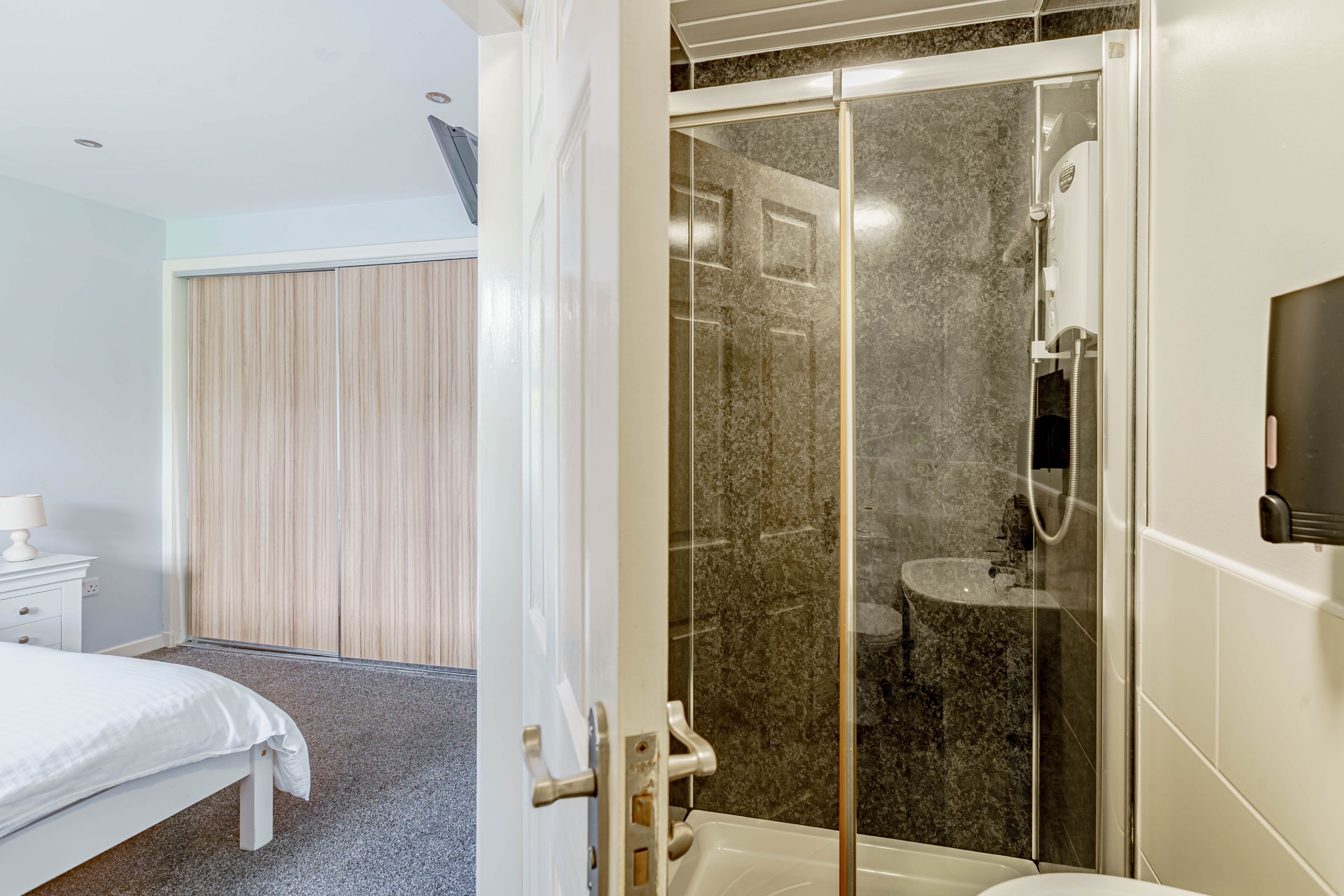 Hotellihuone, jossa on sänky, yöpöytä, lasiovilla varustettu suihku ja wc.
