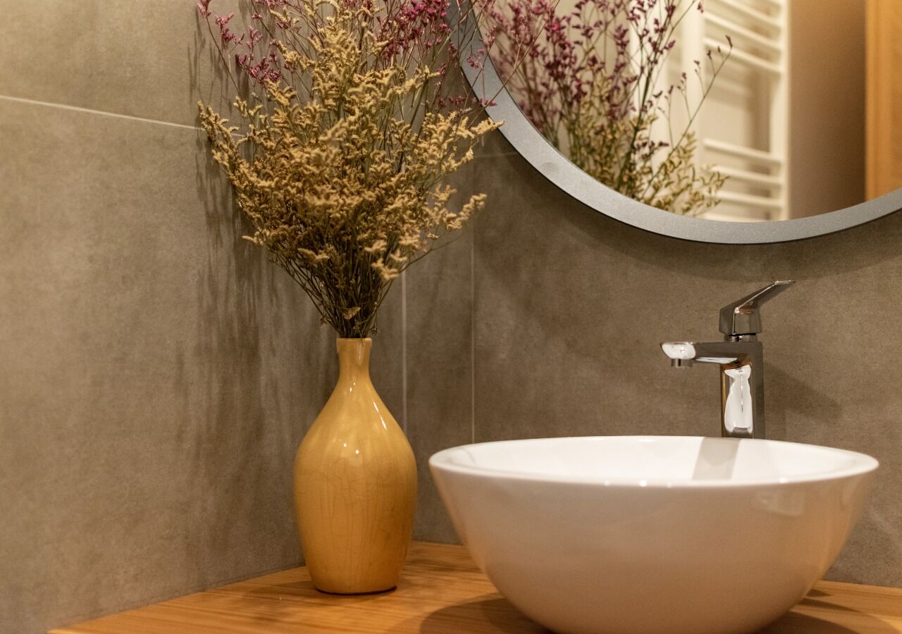 A bathroom with a white sink, a vase with dried flowers, and a mirror reflecting the flowers.