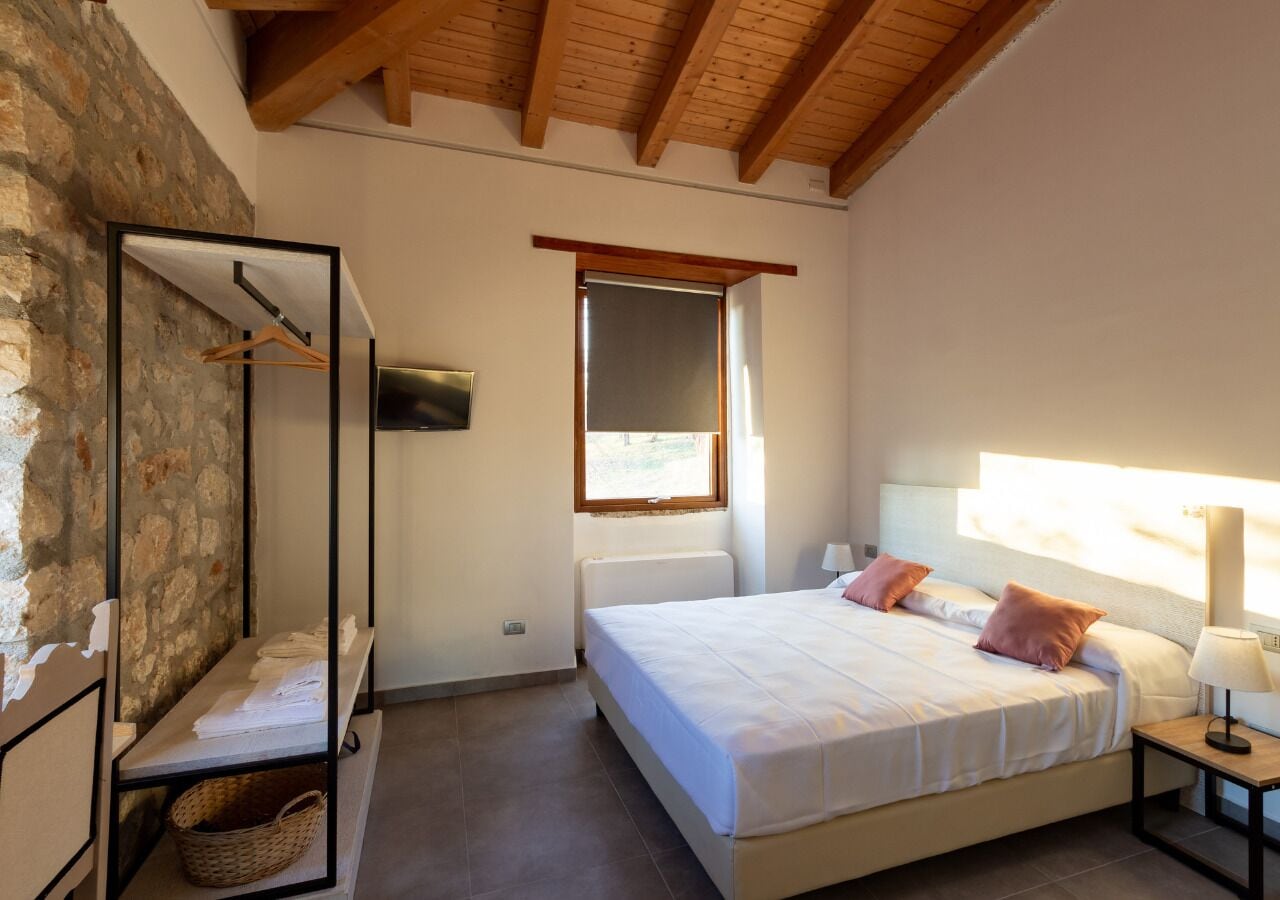 A bedroom with a large bed, a wooden ceiling, a stone wall, and a window.
