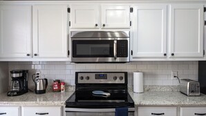 Una cocina con armarios blancos, un microondas de acero inoxidable, una estufa negra, una cafetera, un tostador y un dispensador de toallas de papel.