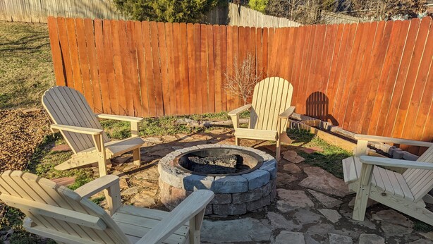 Un patio trasero con un hogar a leña de piedra rodeado por cuatro sillas Adirondack de madera y una mesa de madera.